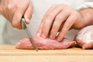 Chef's hands with a knife cutting a meat on the wooden board in the kitchen. Preparation for cooking. Food concept.