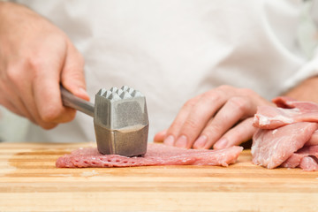 Chef's hands with a hammer beating a meat on the wooden board in the kitchen. Preparation for cooking. Food concept.