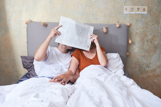 Couple in bed, holding hands, holding newspaper in front of faces