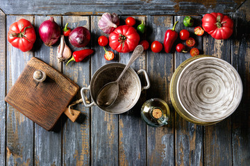 Variety of raw organic vegetables tomatoes, pepper, garlic, onion with empty bowl, pan, olive oil, cutting board over old wooden plank background. Top view with copy space.