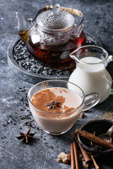 Glass cup of traditional indian masala chai tea with ingredients above. Cinnamon, cardamom, anise, sugar, black tea in glass teapot, jug of milk over dark texture background with space