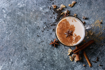 Glass cup of traditional indian masala chai tea with ingredients above. Cinnamon, cardamom, anise, sugar, black tea over dark texture background. Top view with copy space