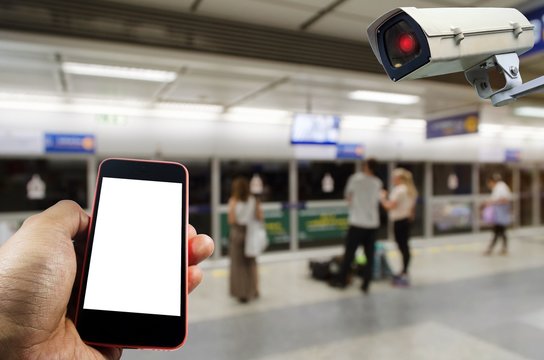 hand using smart phone and CCTV security camera system operating with blurred image of people waiting for subway at train station, internet network, surveillance security, safety technology concept