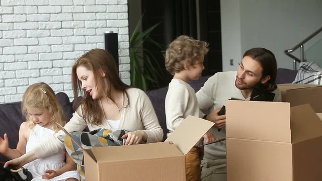 Smiling family with small son and daughter unpacking boxes together sitting on couch in modern cozy living room, happy parents playing with kids unboxing belongings after relocation into new home