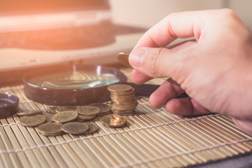 The abstract image of the hand put a coin on the coins stack on the table laptop computer and magnifier is backdrop.