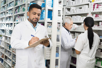 Chemist Writing On Clipboard While Colleagues Standing By Shelve