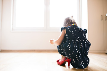 Unrecognizable little girl in dress and red high heels at home.