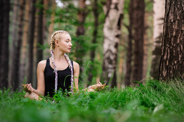 A beautiful slender girl with long braids meditates standing near an old pine tree with outgrowths on the trunk