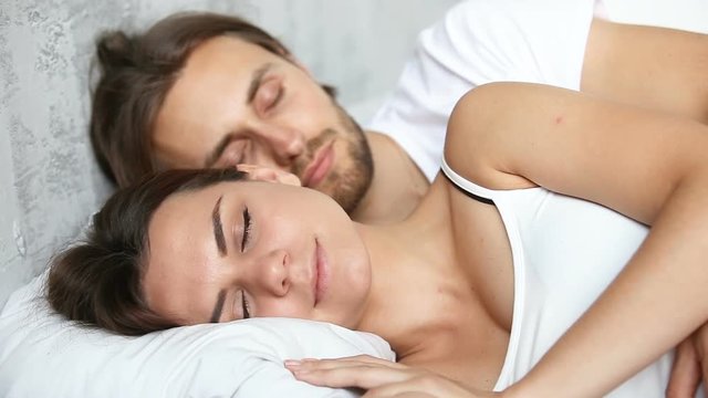 Young couple sleeping in bed, loving husband embracing wife lying on white linen with eyes closed enjoying smiling in light nap in the morning before waking up, happy woman and man resting together