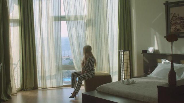 Beautiful young woman in the striped trouser suit sitting on the ottoman in the hotel room