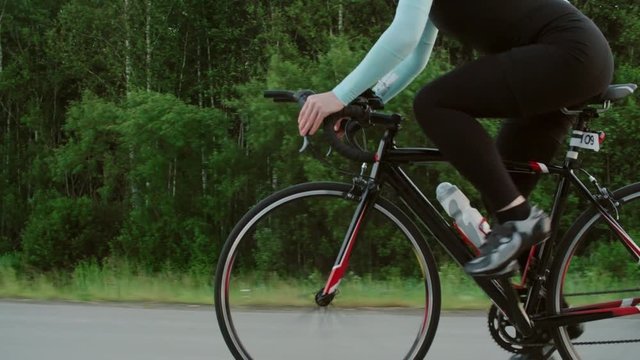 Tracking with tilt down of female cyclist in sunglasses and protective helmet pedaling bicycle along road surrounded by lush green forest