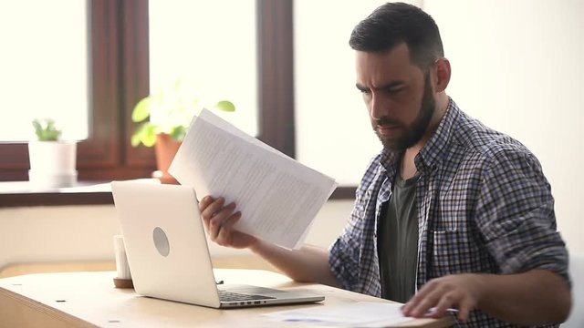 Stressed unmotivated man confused by mistake in documents, looking through papers, frowning using laptop, failing urgent task, missing deadline, quits after bad work fed up with difficult job