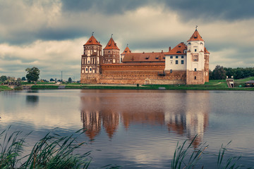 Naklejka na ściany i meble View of Mir Castle - ancient medieval fortress on the shore of the lake, reflecting in the water. Dramatic cloudy sky. Architectural monument of the Belarus. UNESCO World Heritage. Vintage effect. 