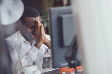 Tired African man sitting at a office after a hard workday, working on laptop, trying to concentrate