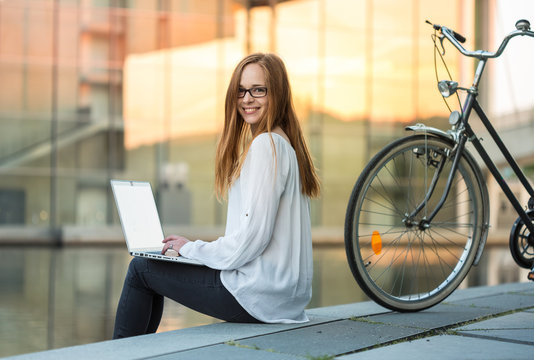 a young woman sits near the river somewhere in the city, works with a laptop with smile; her bicycle is standing next to her; warm golden sunlight is reflected from glass buildings across the river