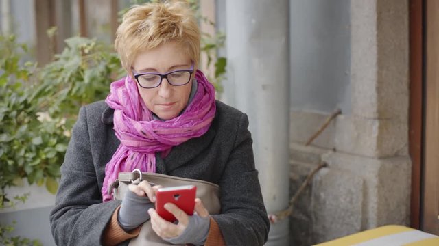 Adult blonde woman browsing smartphone and having drink from paper cup in outside cafe.