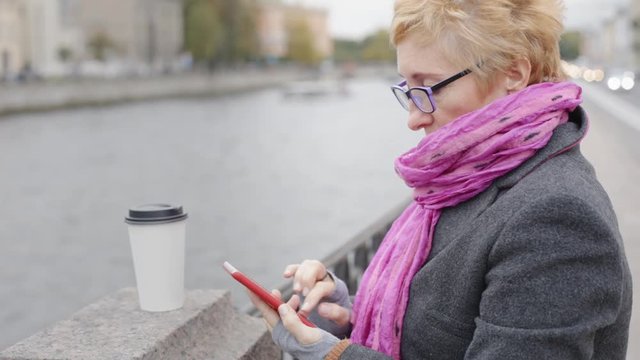 Adult woman in glasses standing with smartphone and coffee on waterfront.