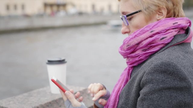 Adult woman in glasses standing with smartphone and coffee on waterfront.