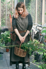 Young cute lady working at the greenhouse holding basket with wildflowers and wearing linen dress and leather belt with florist tools