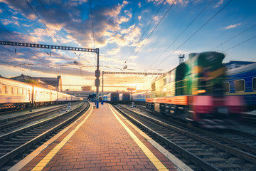 Amazing industrial view with old locomotive in motion, passenger trains with blue wagons, railway station and colorful sky with clouds at sunset. Blurred train on railroad. Beautiful railway platform