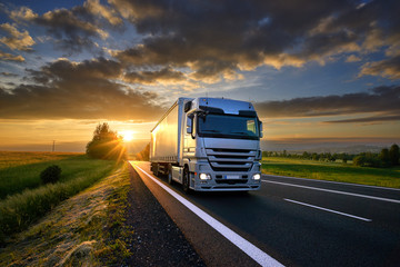 Truck driving on the asphalt road in rural landscape at sunset with dark clouds - obrazy, fototapety, plakaty