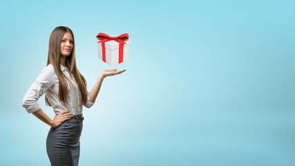 A young longhaired businesswoman stands with one palm up supporting a hovering gift box.