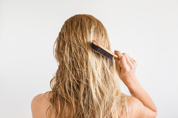 Woman with comb brushing her wet blonde hair after shower on the gray background. Cares about a...