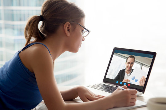 Woman in glasses watching business webinar and taking notes. Aspiring female entrepreneur watching free video lesson with businessman showing potential profits for startup business on laptop screen.