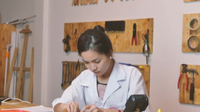 Medium shot tilt down to Asian  female youth testing an Arduino controller board in a makerlab.  The words 'Maker Tools' and many tools and instruments can be seen on the wall behind her.