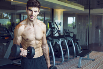 Young handsome muscular man exercising in the gym. Dumbbell bicep curl exercise in standing position next to the weight rack. Exercise with the smile.