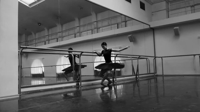 Ballerina in black tutu and pointe stretches on barre in ballet gym. Woman standing near bar and mirror, preparing for perfomance. Slow motion. Black and white tonned footage