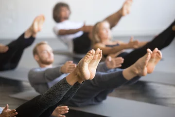 Fotobehang Group of young sporty people practicing yoga lesson with instructor, stretching in Paripurna Navasana exercise, balance pose, working out, indoor close up image, studio, focus on feet © fizkes