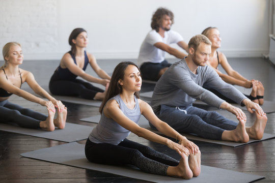 Group of young sporty people practicing yoga lesson with instructor, sitting in paschimottanasana exercise, Seated forward bend pose, indoor full length, studio, friends working out in club