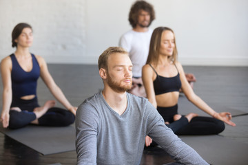 Fototapeta na wymiar Group of young people, sporty students practicing yoga lesson with instructor, sitting and meditating with closed eyes in Padmasana exercise, Lotus pose, friends working out in club, studio