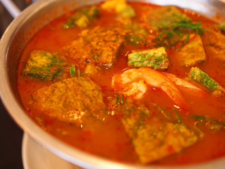 Red orange Thai local traditional hot sour spicy curry soup, “Kang Som”, with deep fried omelet with green leaf, “Cha Om”, and shrimps, in stainless steel round bowl, close up shot
