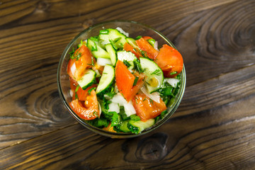 Fresh salad with tomato, cucumber, onion, parsley and dill in glass bowl on wooden table