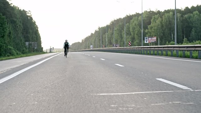 Dolly with wide shot of female cyclist in professional sportswear and protective helmet riding bicycle along highway surrounded by green forest