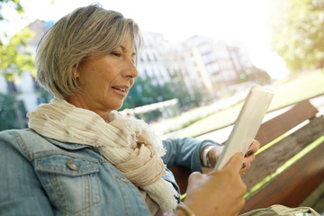 Senior woman sitting on public bench connected with tablet