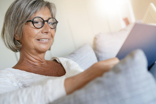 Senior woman with eyeglasses reading book in sofa
