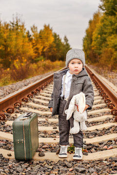 a lost little boy in vintage clothes hugs a bunny next to an old-fashioned suitcase in the middle of the forest on an abandoned railway rails