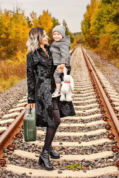 Mother holds her son on the arms with a suitcase on the train tracks in the middle of autumn forest