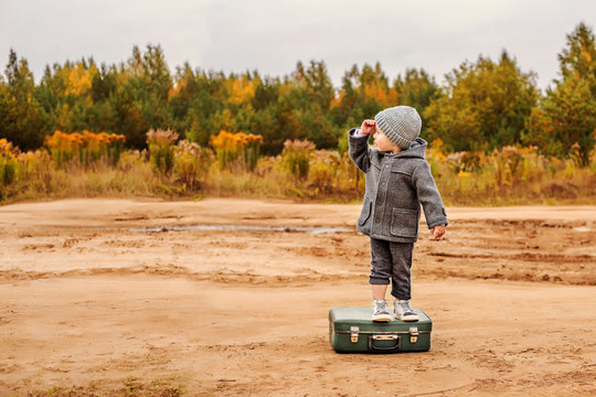 a boy in panties and a gray coat is standing on an old suitcase in the middle of a sandy country road, raising his hand to his forehead