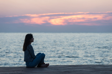 Lonely girl sitting on the sunrise beach. Woman silhouette over sunrise sky