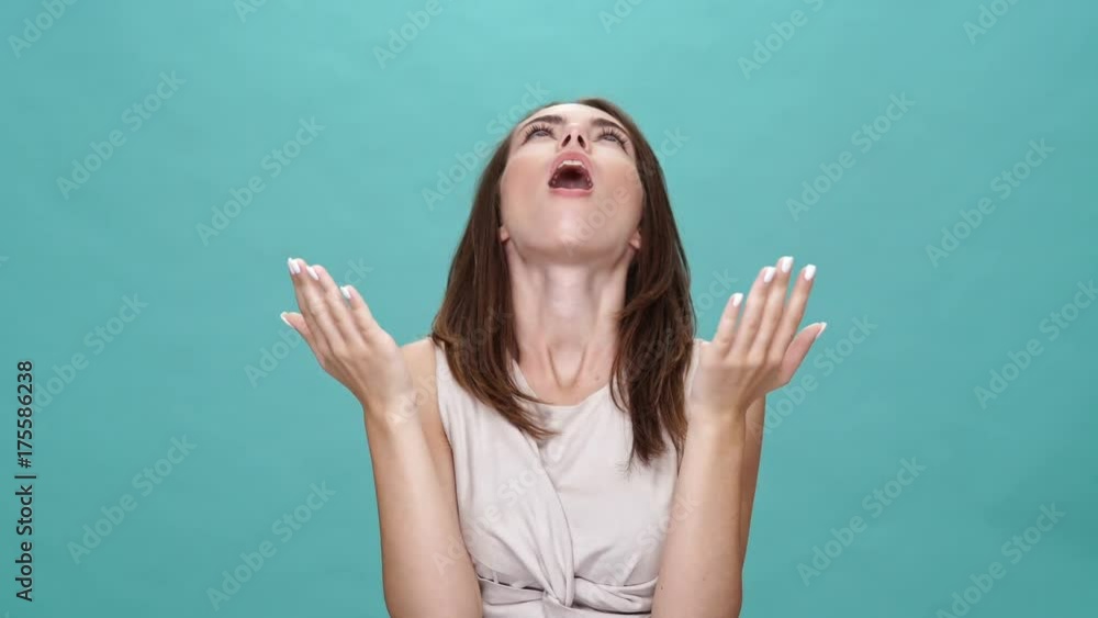 Poster shocked happy brunette woman in t-shirt looking up and at the camera over turquoise background