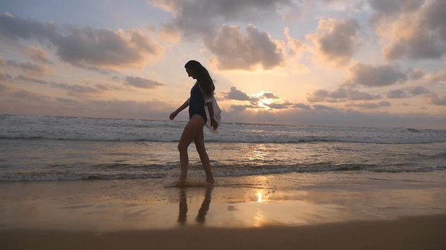 Happy woman walking and spinning on the beach near the ocean. Young beautiful girl enjoying life and having fun at sea shore. Summer vacation or holiday. Sunset landscape at background. Slow motion