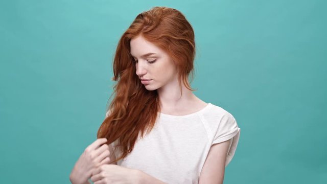 Smiling ginger woman in t-shirt touching her hair and looking around over turquoise background