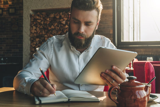 Young bearded businessman sits in cafe, home at table, holding tablet computer and writes in notebook. Man is working, student is studying. Online education, marketing, training.E-learning,e-commerce.