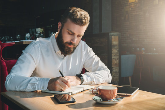 Young bearded businessman sits in cafe, home at table and writes in notebook. On table tablet computer, smartphone.Man is working, planning.Online education,marketing, training.E-learning, e-commerce.