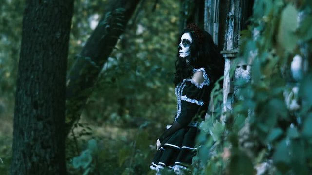 The Mexican Day of the Dead. The young woman with spooky sceleton make-up for Halloween dressed in black clothes leaning to wooden wall and looking at camera. 4K
