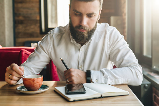 Young bearded businessman, dressed in white shirt, sits in cafe, office at table, holds pen in his hand, reads information on smartphone screen, drinks coffee. On desk is notebook. Online education.